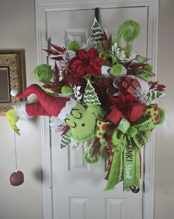 A Christmas wreath with red poinsettias, small green trees and a Grinch peeping out of the middle.