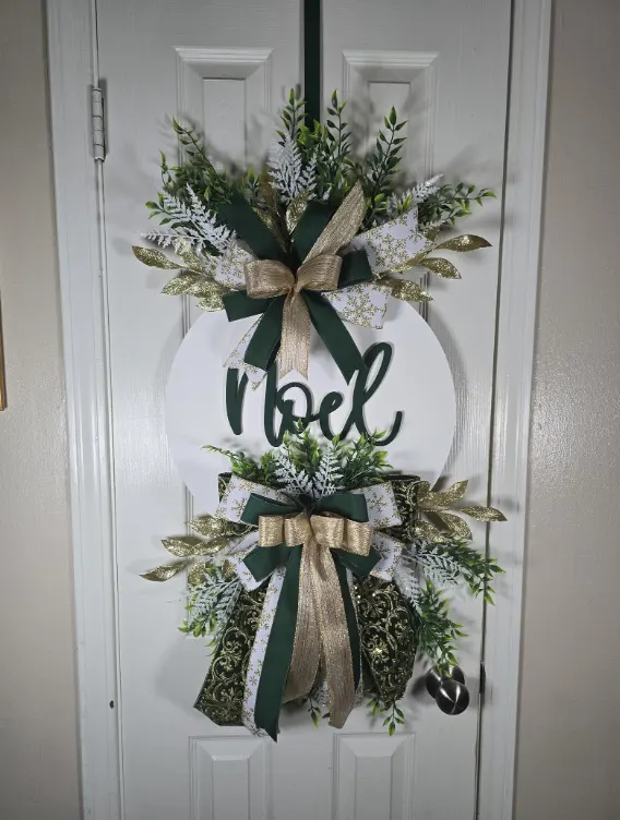 A white hoop wreath reading Noel, with frosted greenery and a deep green velvet bow.
